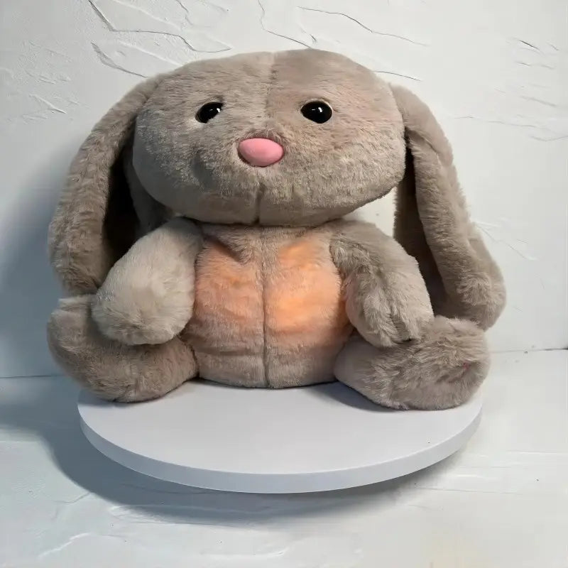Ein Plüschkaninchen aus hellem Grau, das atmet; ein beruhigendes Sinnes-Plüschbabyspielzeug mit langen, herabhängenden Ohren und einer weichen rosafarbenen Nase; sitzt auf einem weißen runden Sockel.