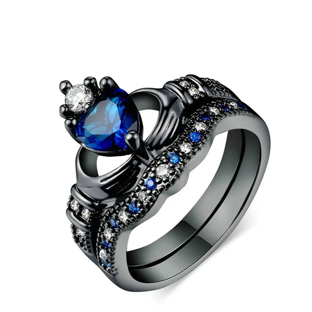 Black titanium claddagh wedding ring set featuring a prominent blue heart sapphire stone and smaller clear stones with intricate detailing.