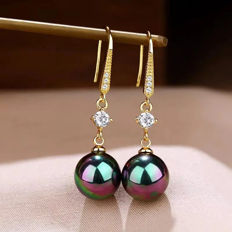 Elegant pearl and pave diamond drop earrings in gold tone featuring a row of sparkling diamonds along the hook and a large black pearl with iridescent green and purple hues.