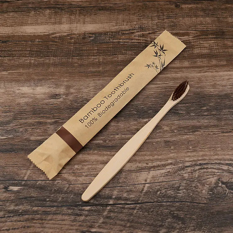 Un cepillo de dientes de bambú natural con cerdas suaves de color marrón oscuro, junto a su envoltorio de papel respetuoso con el medio ambiente de un juego de 10 unidades de cepillos de bambú.