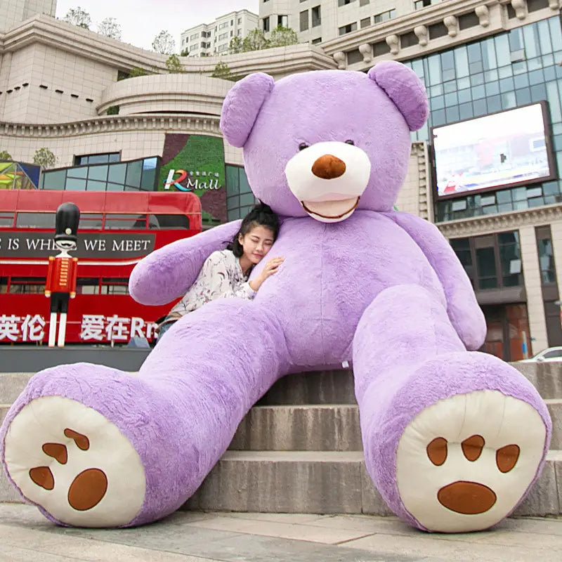 Ein flauschiger, lavendelfarbener Riesen-Teddybär mit weißen Pfoten und brauner Nase, der auf städtischen Stufen aus weichem Plüschmaterial sitzt, mit einer Größe zwischen 1 m und 3,4 m.