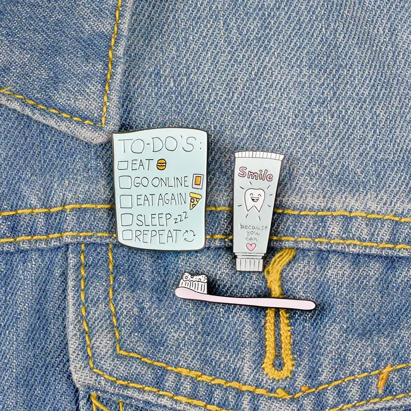 A light blue enamel pin shaped like a to-do list with checkboxes and food icons accompanied by matching toothpaste and toothbrush cartoon enamel pins.
