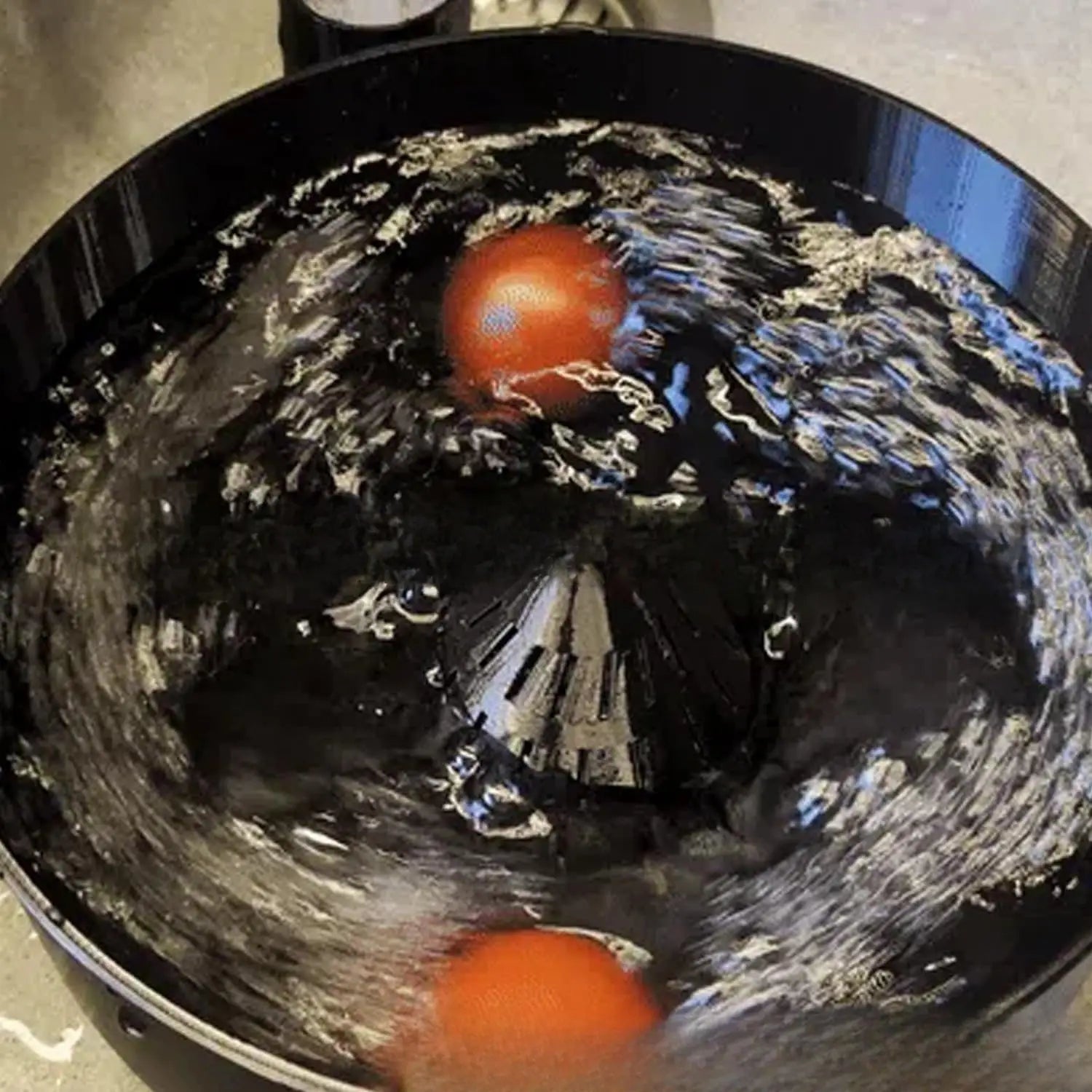 Une tomate rouge flottant dans un bol de lavage de fruits noir en plastique durable, avec de l’eau tourbillonnante, faisant partie d’un ensemble de bol de lavage de fruits et de presse-agrumes.