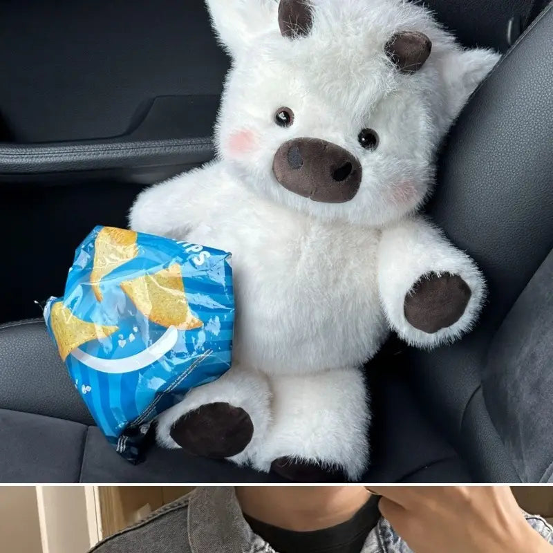 white plush cow soft stuffed animal toy with black nose and hooves and pink cheeks sitting in a car seat holding a blue bag of chips for kids