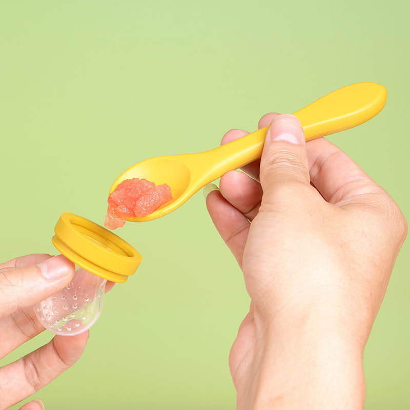 Gelber Kunststoff-Babylöffel zum Füttern mit einer Portion rosa Püree, der von einem transparenten Silikon-Füttereinsatz in einem Baby-Obst-Füttergerät aus Silikon, Teether und Schnuller-Set übertragen wird.
