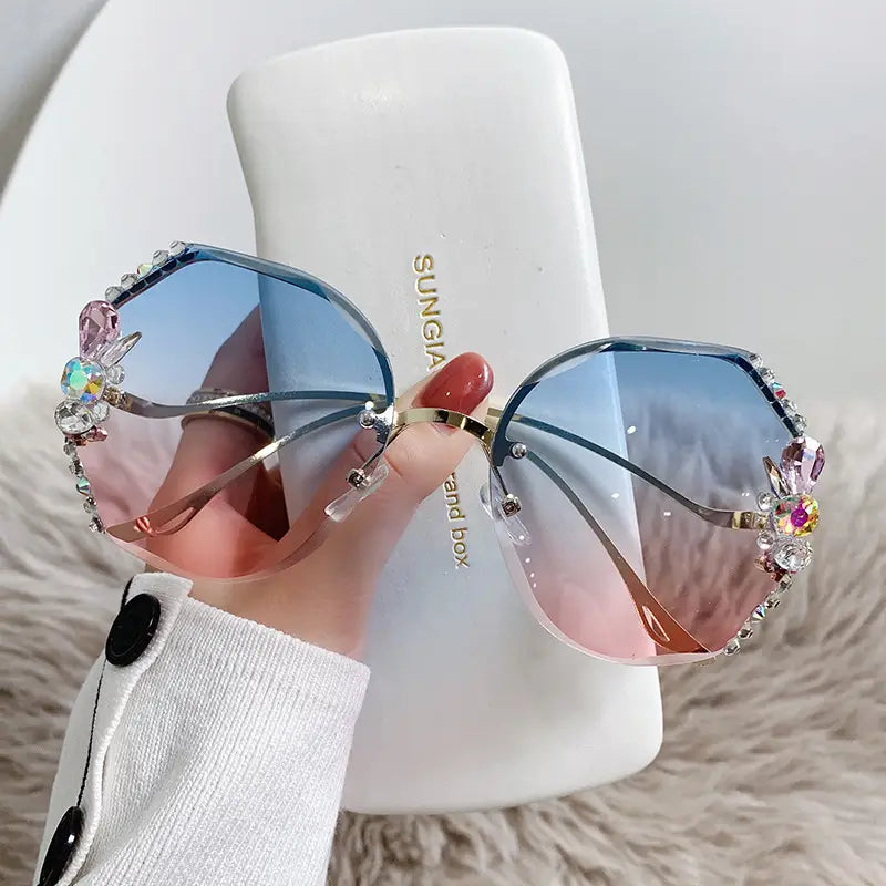 Un par de gafas de sol de lujo con pedrería de mariposa tipo cristal y protección UV400, con monturas extragrandes y lentes con degradado azul a rosa, así como pedrería ornamentada que brilla y adornos de cristal con gemas de colores.