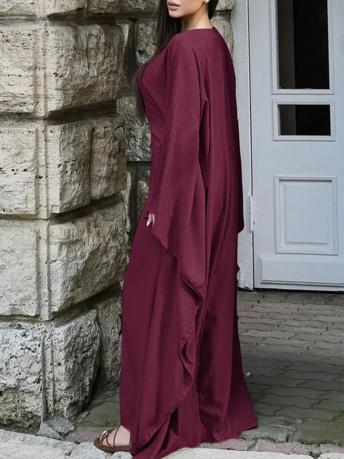 A flowing maroon satin maxi dress with a draped asymmetrical design, long sleeves, and a ruffled hem.