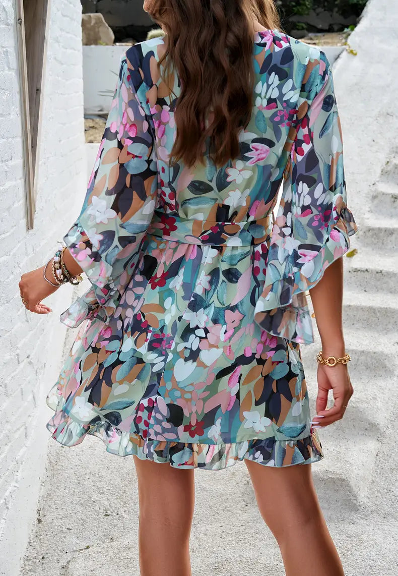 A flowing floral v-neck mini dress with ruffled sleeves and hem in soft pastel tones featuring a waist tie.