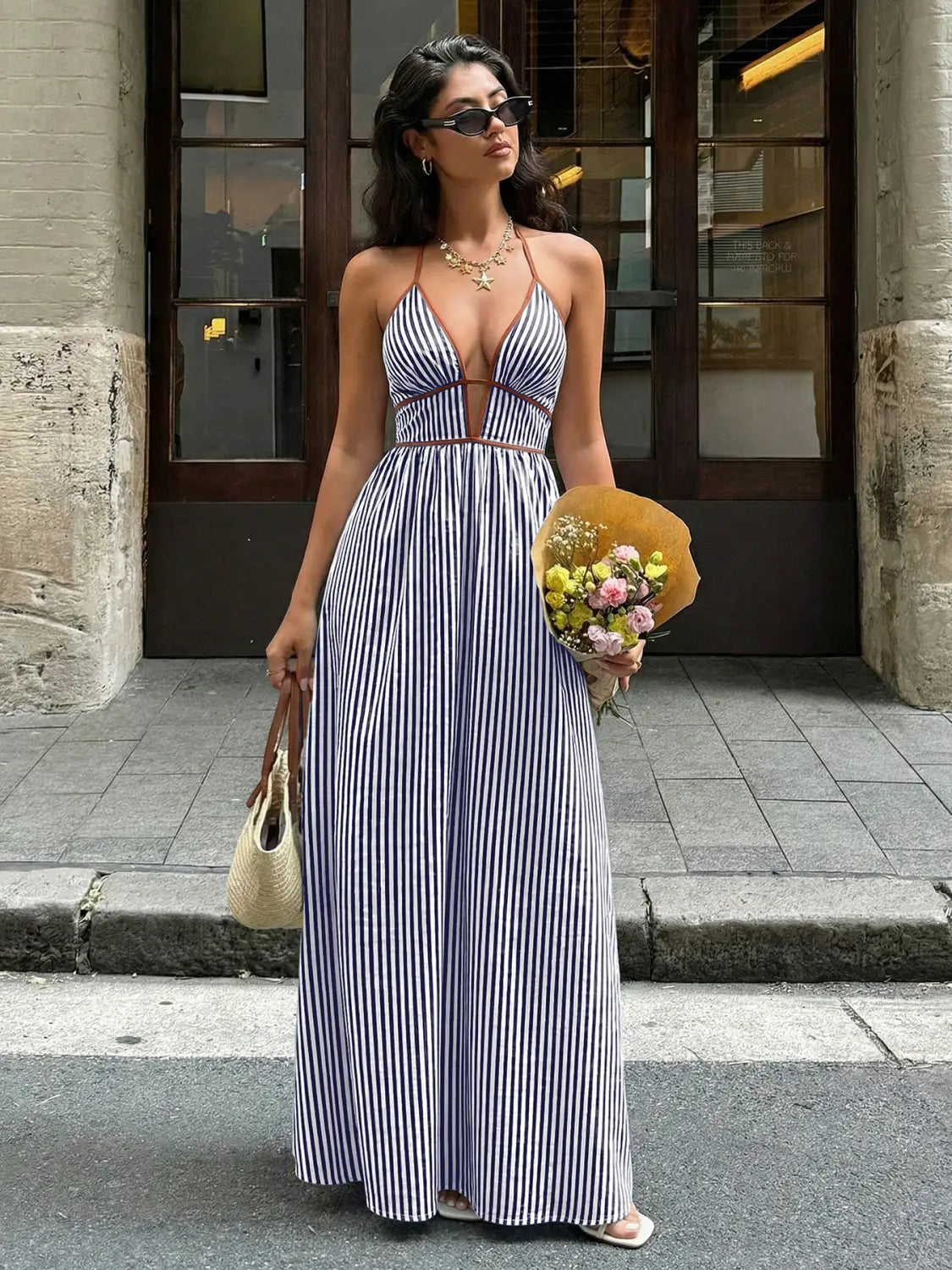 Une femme porte une robe longue d’été maxi à encolure en V et dos noué à rayures, bleu marine et blanche, à rayures verticales, avec une encolure type halter et un détail découpé au niveau de la poitrine.