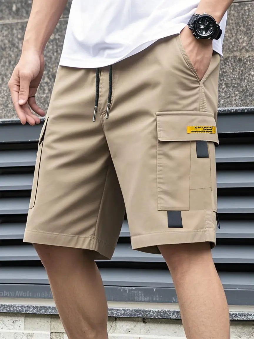 Shorts cargo utilitaires pour homme en beige, avec un cordon noir, une étiquette jaune sur la poche de la cuisse et des accents gris foncé, conçus pour une tenue d’été décontractée.