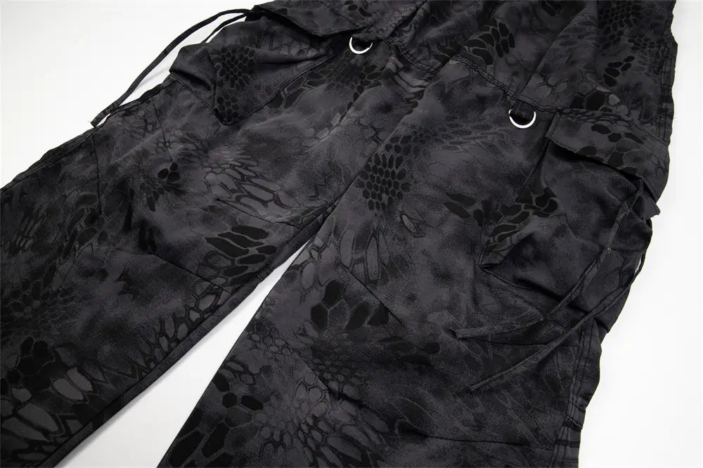 Black tactical cargo pants with a subtle dark-toned snake-scale camouflage pattern, reinforced stitching, and metal d-rings on the pockets designed for durability and wide leg comfort.