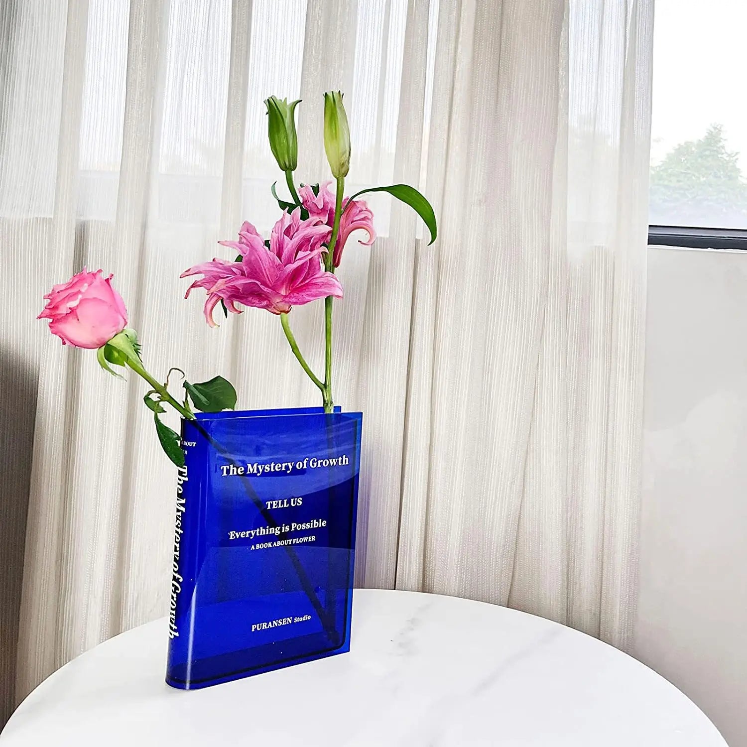 un jarrón de acrílico azul translúcido con forma de libro titulado El misterio del crecimiento que contiene lirios rosados y una rosa para decoración moderna del hogar