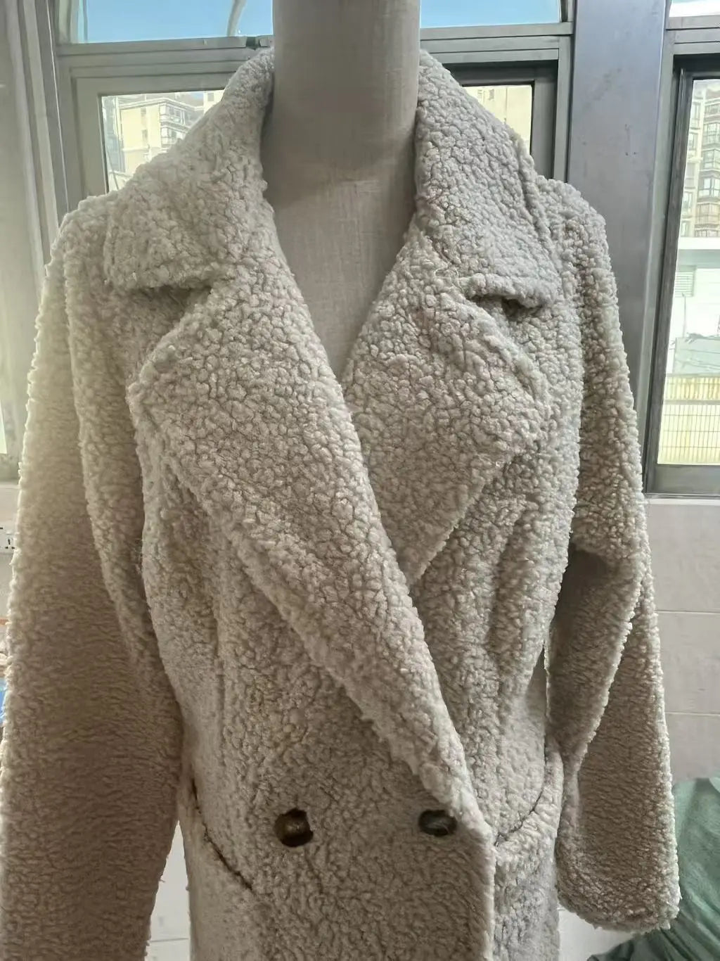 Manteau teddy mi-long doublé en polaire de couleur crème, avec une texture moelleuse et une fermeture à double boutonnage à l’avant, munie de boutons marron foncé.