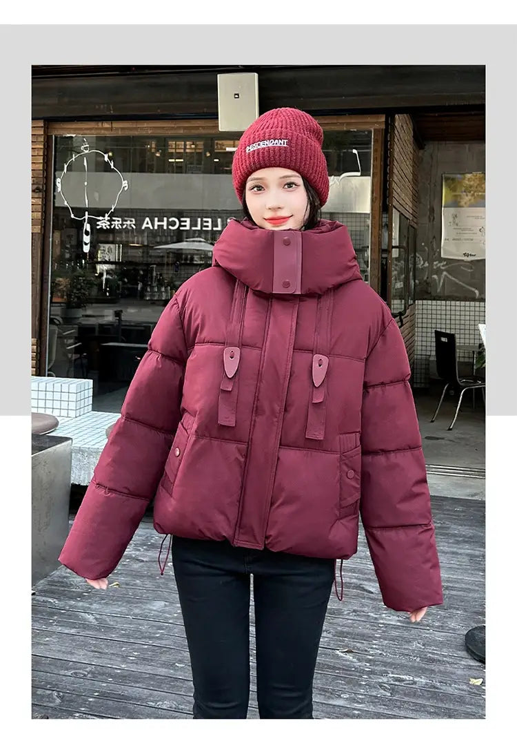Veste courte matelassée à capuche pour femme bordeaux, avec col haut, fermeture éclair frontale et deux poches à rabat avec passants assortis, conçue pour l’habillement d’hiver chaud.