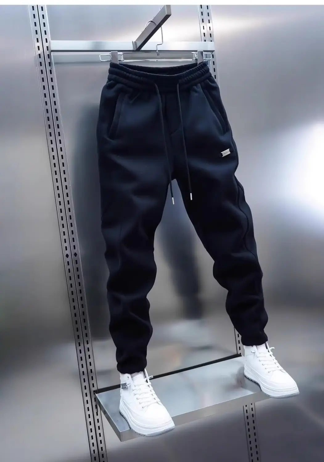 Black mens casual solid color sweatpants with a drawstring waistband and subtle white logo on the thigh hanging on a metal rack.
