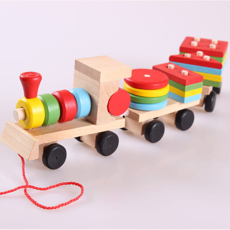 Jouet éducatif en bois de type train pour empiler des formes, avec une structure en bois naturel, une poignée rouge, des roues noires, des anneaux d’empilage aux couleurs de l’arc-en-ciel et des blocs de formes géométriques disposés en couches empilées.