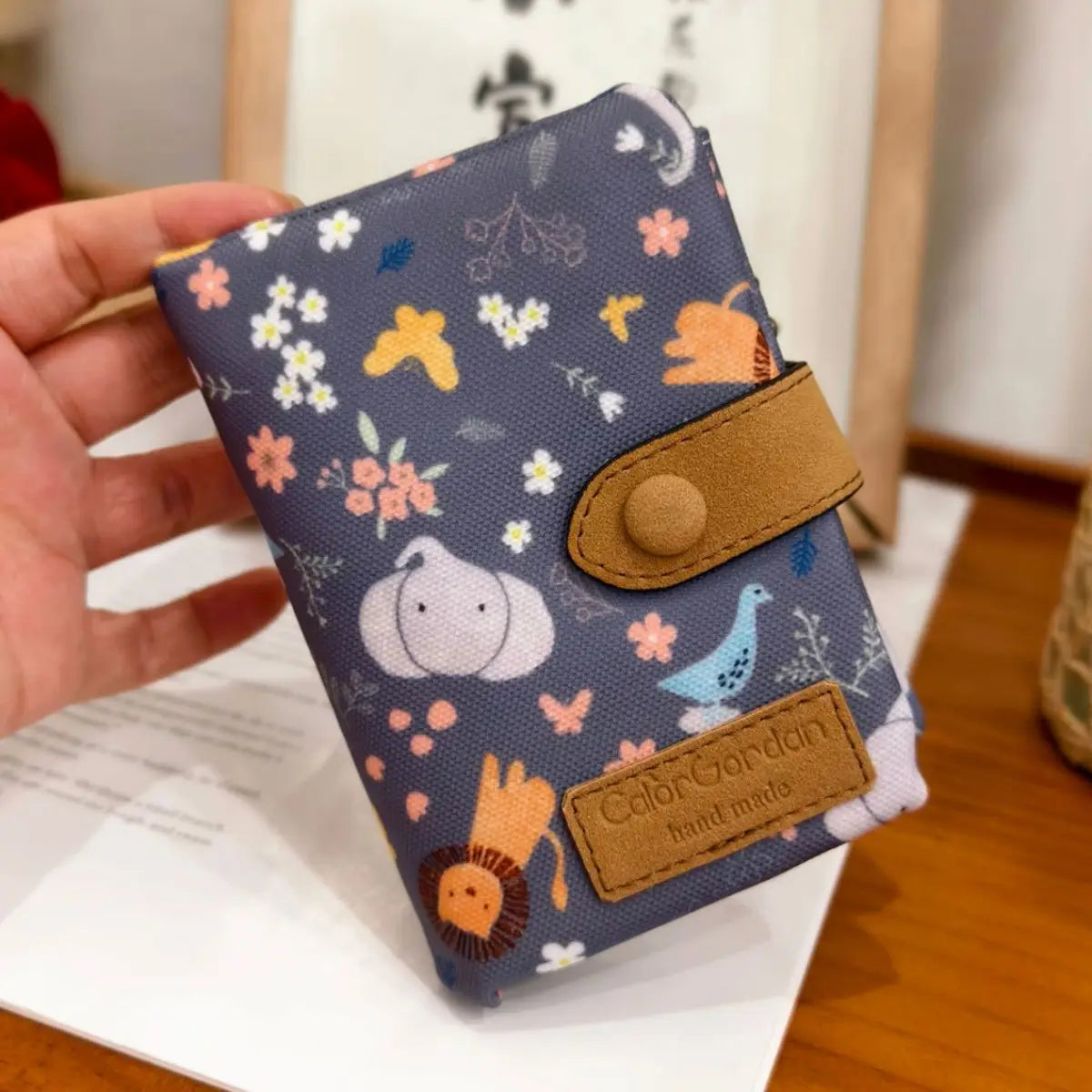 A navy blue canvas tri fold multi card wallet featuring a whimsical pattern of cartoon animals and flowers with tan leather straps and a branded patch.