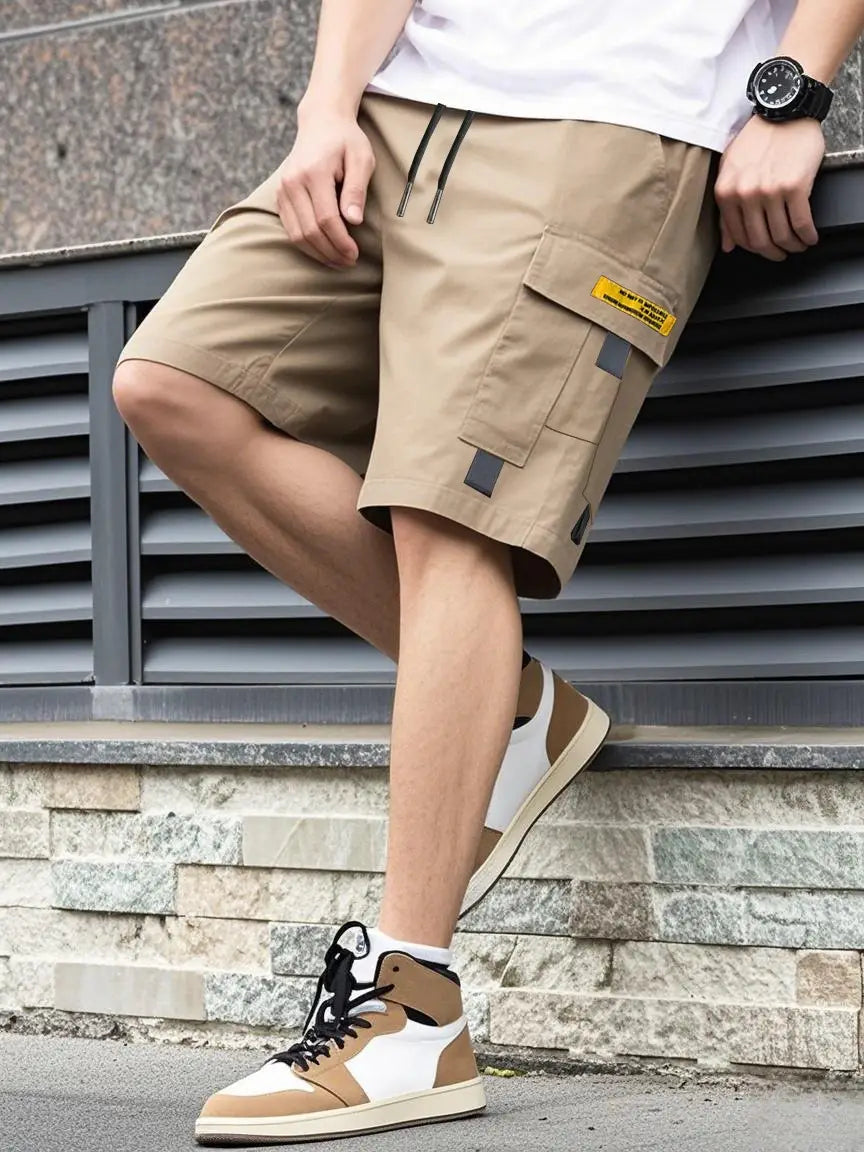 Shorts cargo utilitaires pour homme beige, avec plusieurs poches, cordons noirs et une étiquette jaune sur la poche latérale, conçus pour une tenue décontractée d’été.