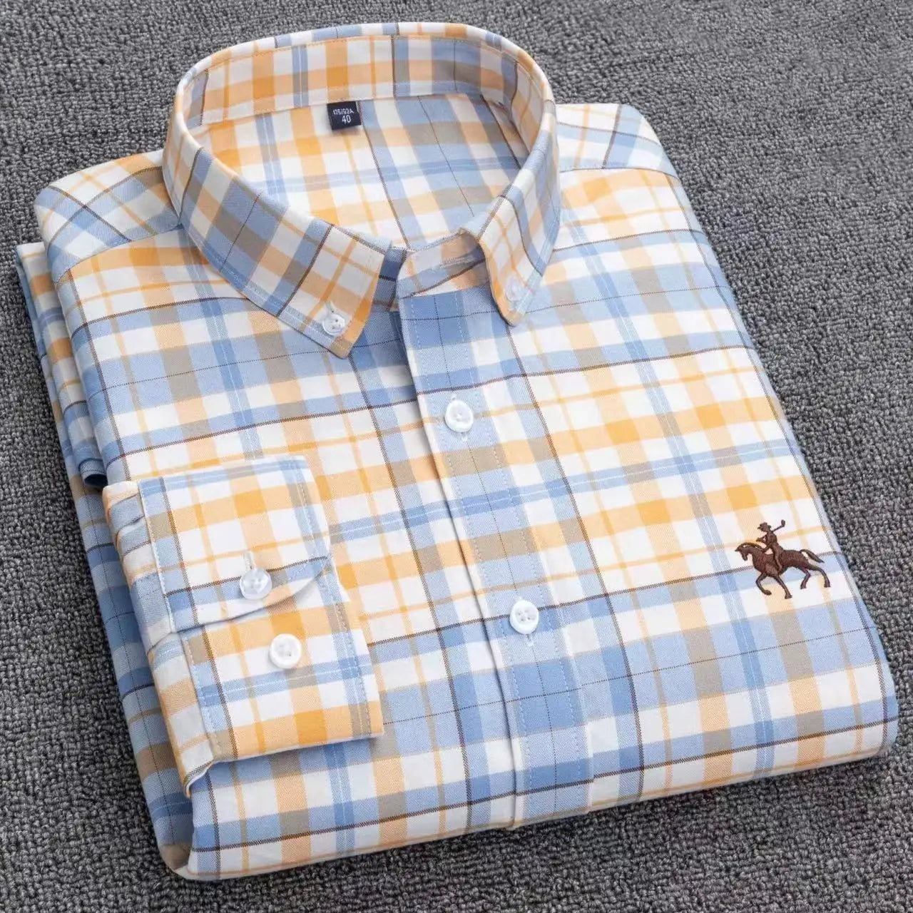 A neatly folded men’s striped cotton embroidered button down shirt in blue yellow and white plaid with a brown embroidered polo player logo on the chest.