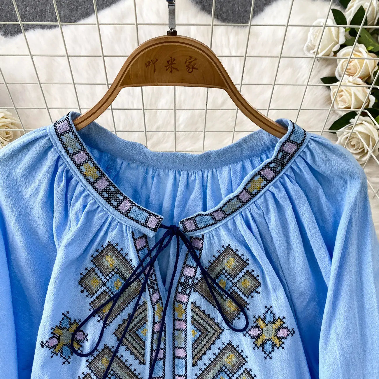 Light blue embroidered lantern sleeve tie neck blouse with soft flowing fabric and intricate geometric pastel pink yellow and beige patterns along the neckline and front.