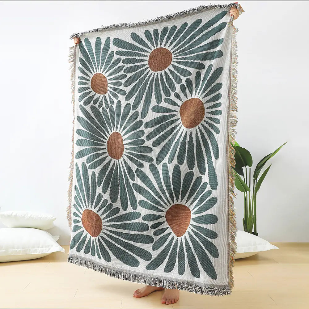 A woven cotton daisy and geometric floral throw blanket with a bold pattern of oversized daisies in teal and tan on a white background framed by textured fringe.
