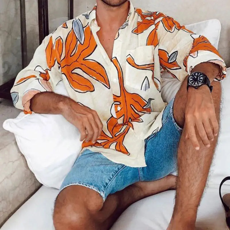 Ein Mann, der ein cremefarbenes Hemd mit langem Ärmel und tropischem Print für Herren am Strand trägt, mit kräftigen Blattmustern in Orange und Grau.