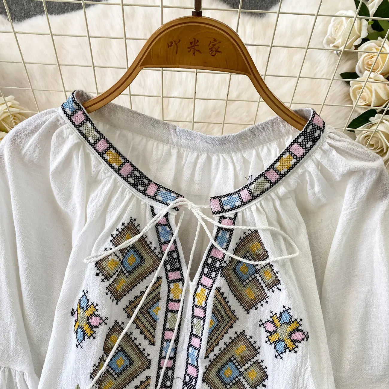 White blouse with intricate geometric pastel pink blue yellow and brown embroidery on the neckline and front panels featuring lantern sleeves and a tie neck design.
