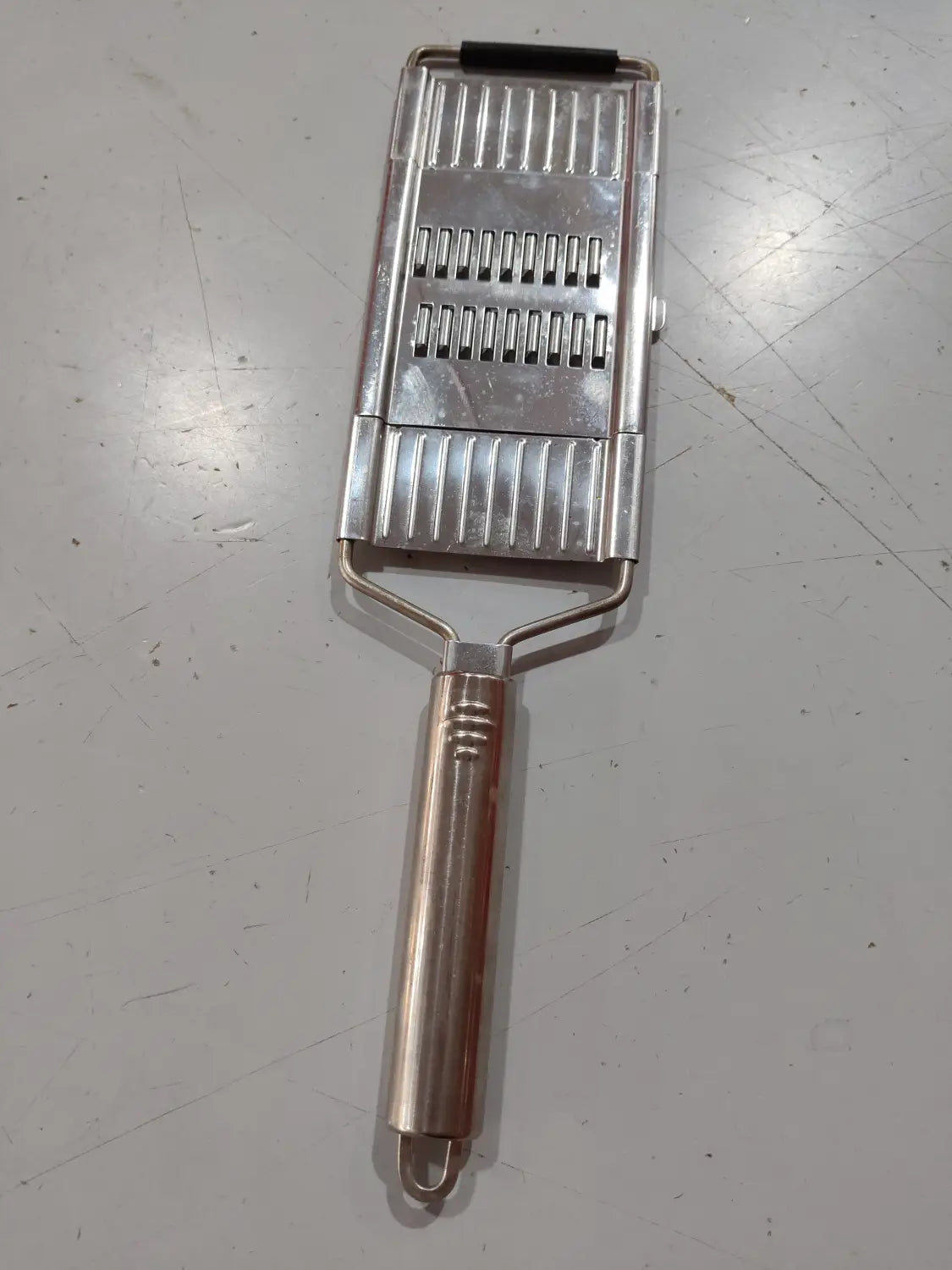 Juego de rallador y cortador multiusos de acero inoxidable con un rallador de verduras metálico plateado con agarre de goma negra en la parte superior y ranuras acanaladas para cortar en láminas.