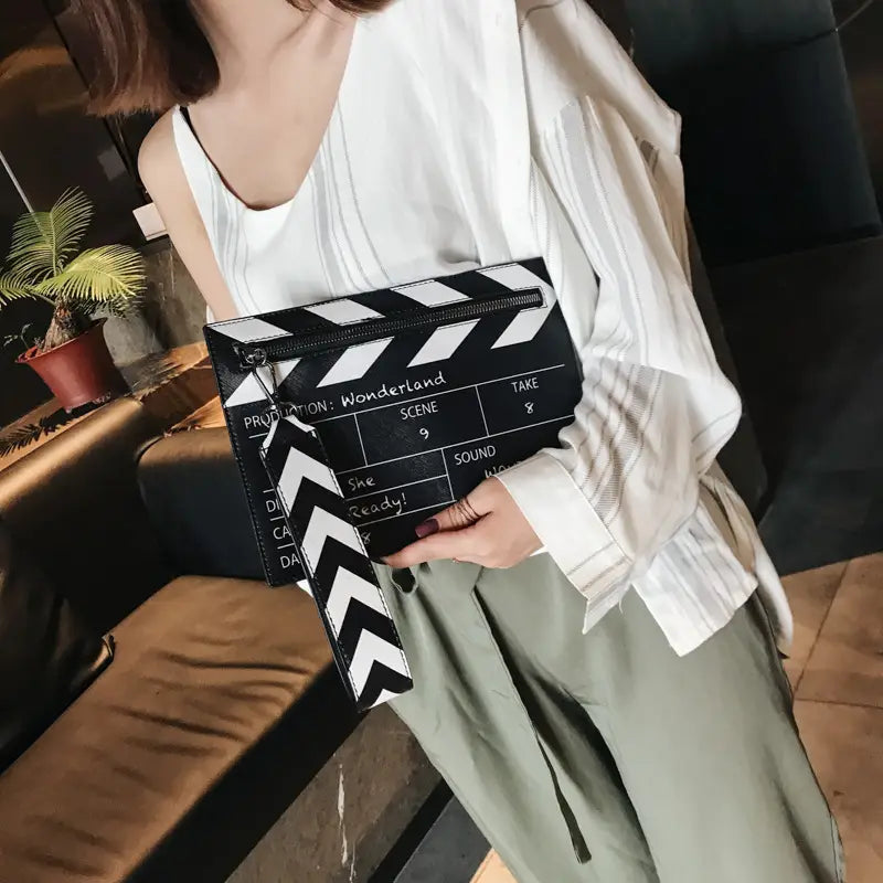 Pochette-sac enveloppe bandoulière en cuir PU noir et blanc, conçue comme un clap de cinéma, avec fermeture à glissière et bracelet de poignet assorti à rayures.