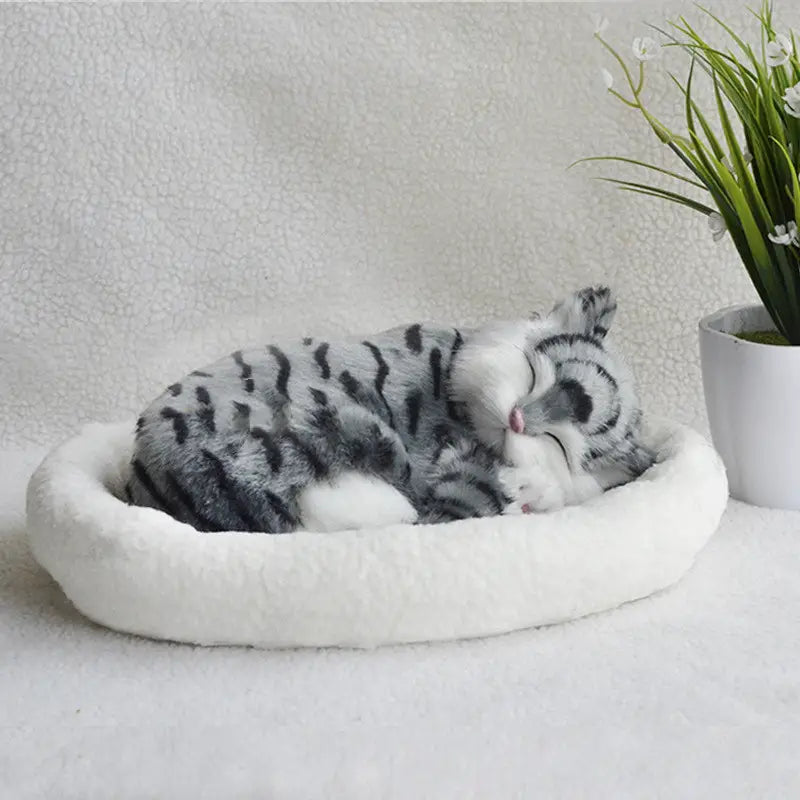 A realistic breathing plush cat figurine in gray and black stripes sleeping peacefully in a soft white circular pet bed.