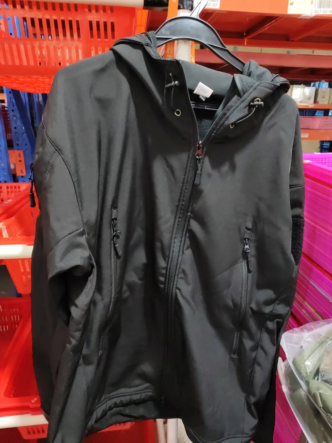 Black hooded mens waterproof tactical fleece soft shell jacket with zippered front and side pockets hanging on a hanger in a retail store setting.
