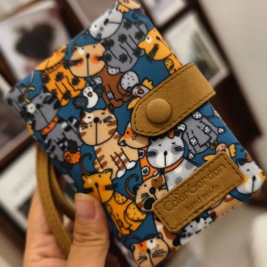 A blue whimsical patterned canvas tri fold multi card wallet featuring a playful pattern of cartoon cats in various colors and designs with a tan leather-like flap and strap.
