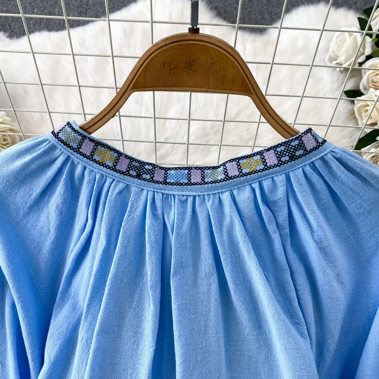 A light blue soft cotton embroidered lantern sleeve tie neck blouse with a decorative pastel floral neckline in lavender and yellow on a dark background.