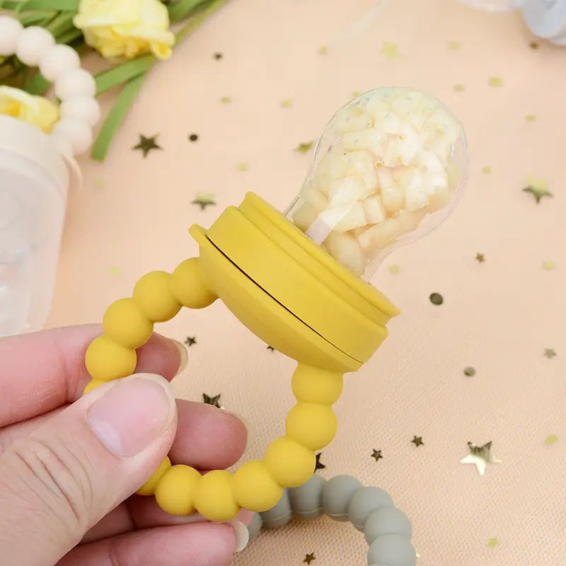 Gelbes Silikon-Baby-Obstfüttergerät, Teether- und Schnuller-Set mit transparentem Kunststoffdeckel, gefüllt mit gewürfeltem Obst, für sicheres Füttern von Säuglingen.