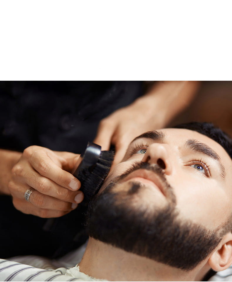 Ein Barbier verwendet eine schwarze, starre Bartschaumbürste mit Schweineborsten und einem Holzgriff, um das Gesichtshaar zu pflegen.