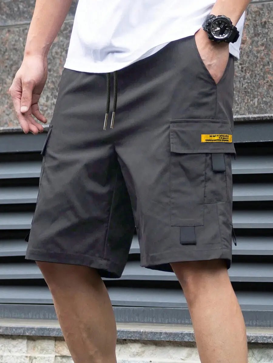 Shorts cargo utilitaires pour homme en gris foncé, avec plusieurs poches et une étiquette jaune sur la cuisse, conçus pour une tenue d’été décontractée, avec une taille à cordon de serrage.