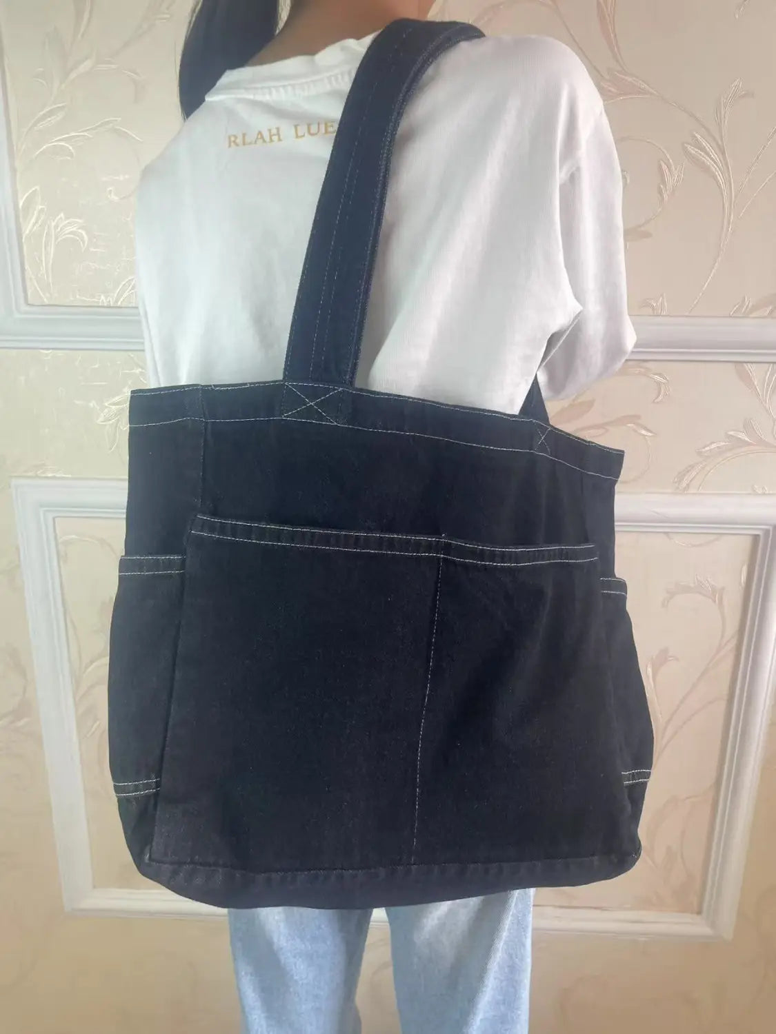 Black denim tote bag with white stitching and two front pockets designed as a large capacity Alma Latina shoulder bag.