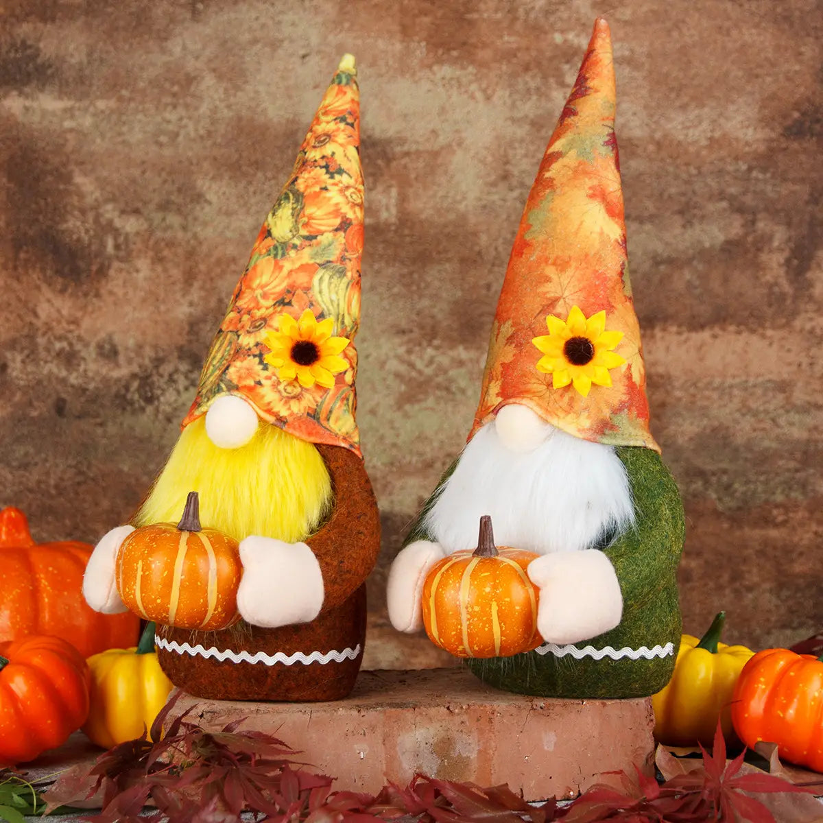 Zwei Danksagungs-Gnom-Plüschfiguren mit Herbstthema: eine mit gelbem Bart und braunem Körper, die einen orangefarbenen Kürbis hält, und eine mit weißem Bart und grünem Körper, die ebenfalls einen orangefarbenen Kürbis hält; beide tragen hohe, spitze Hüte, die mit Mustern aus herbstlicher Vegetation und Sonnenblumen verziert sind.
