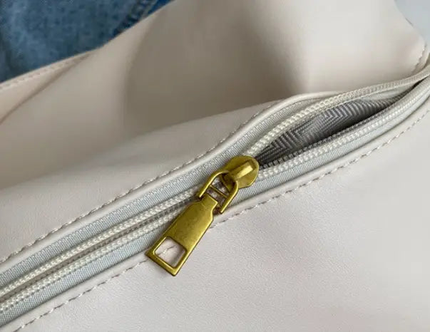 A cream-colored synthetic leather shoulder bag with a gold-toned zipper pull and gold buckle strap.