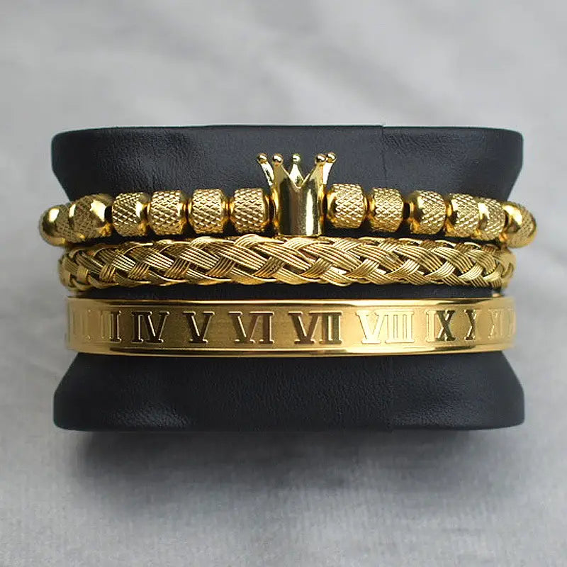 Gold-toned Crown Royal diamond beaded bracelet set featuring a textured beaded band with a central crown charm, an intricate braided design, and a smooth roman numeral engraved bangle.