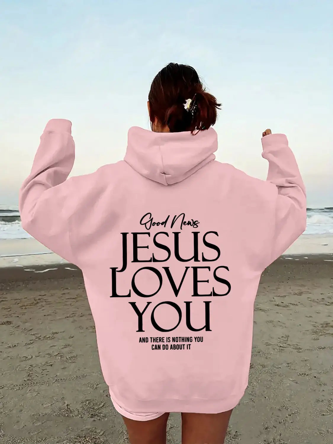 back view of a pink oversized streetwear hoodie with a kangaroo pocket featuring black text reading good news jesus loves you and there is nothing you can do about it across the back.