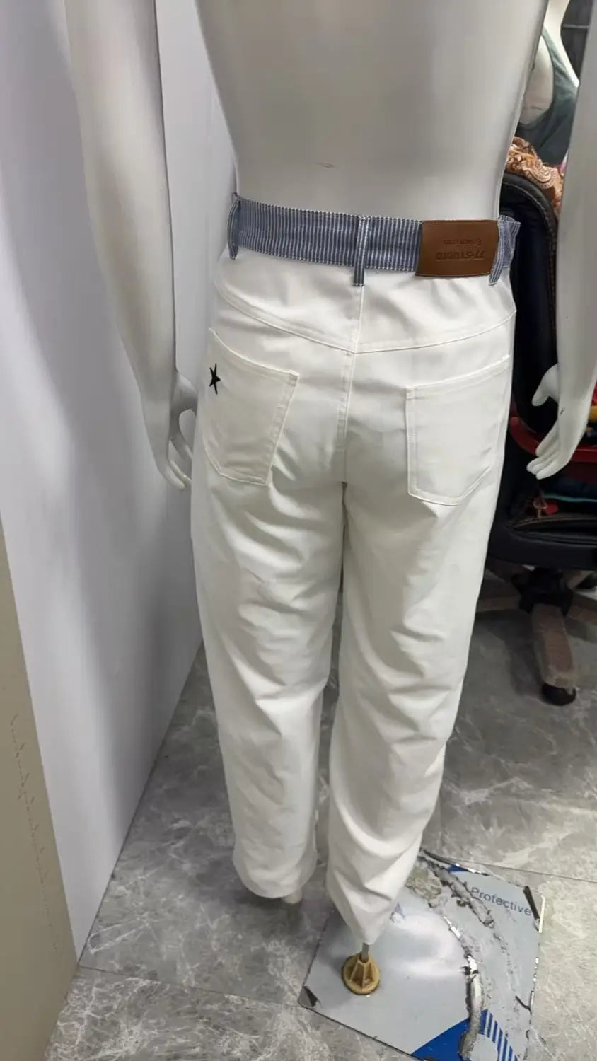 Jean à taille avec bande blanche et jambes évasées présenté de dos, avec tissu en denim blanc, une ceinture rayée bleu et blanc avec une boucle en cuir brun, et une petite broderie d’étoile noire sur la poche arrière.