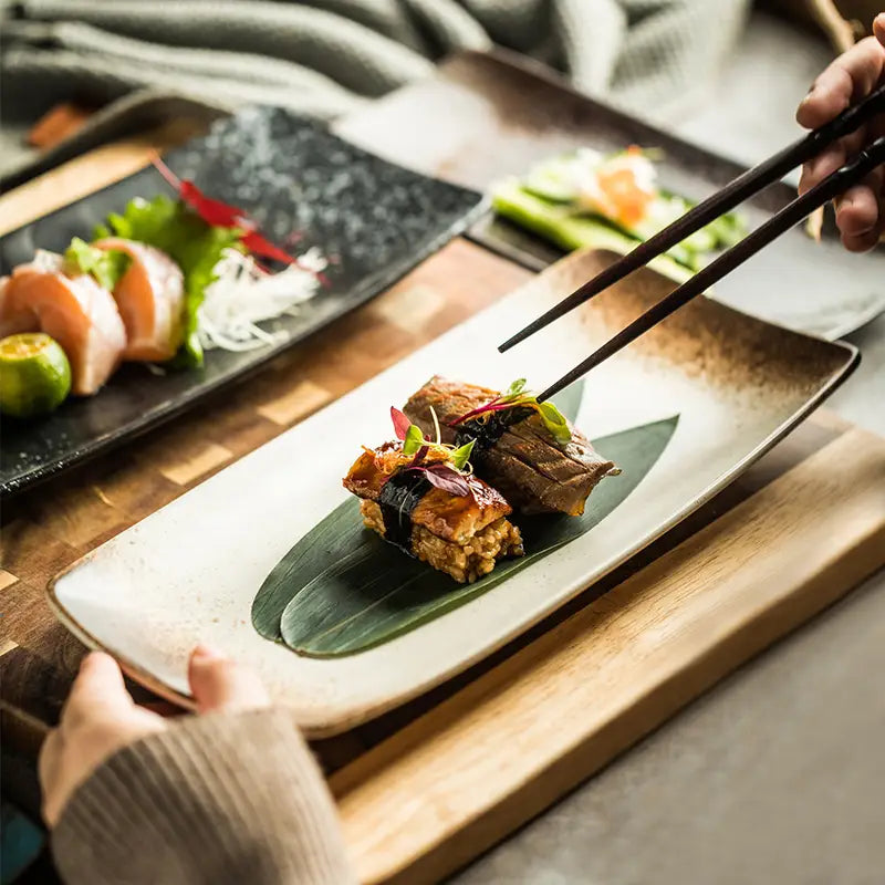 A rectangular Japanese ceramic sushi plate with a matte off-white glaze and subtle speckled texture featuring a single serving of grilled fish garnished on a green leaf.