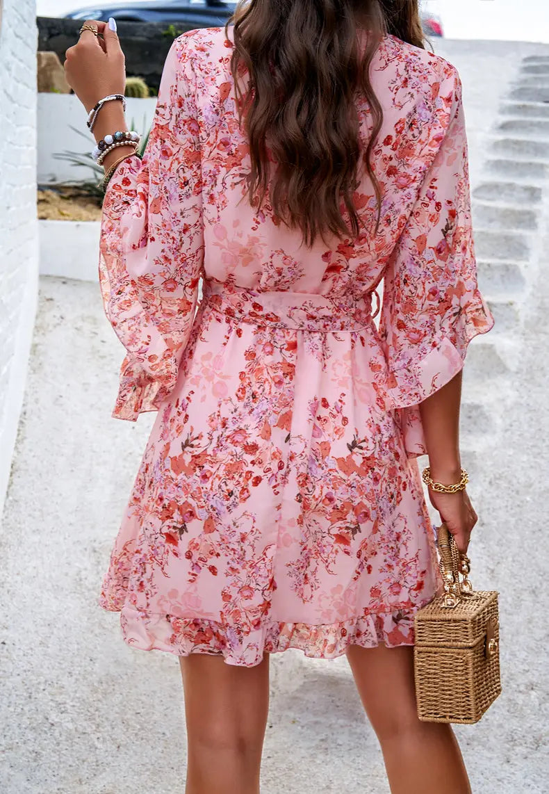 A pink floral print women’s v-neck dress with ruffled sleeves and hem featuring a belted waist tie.