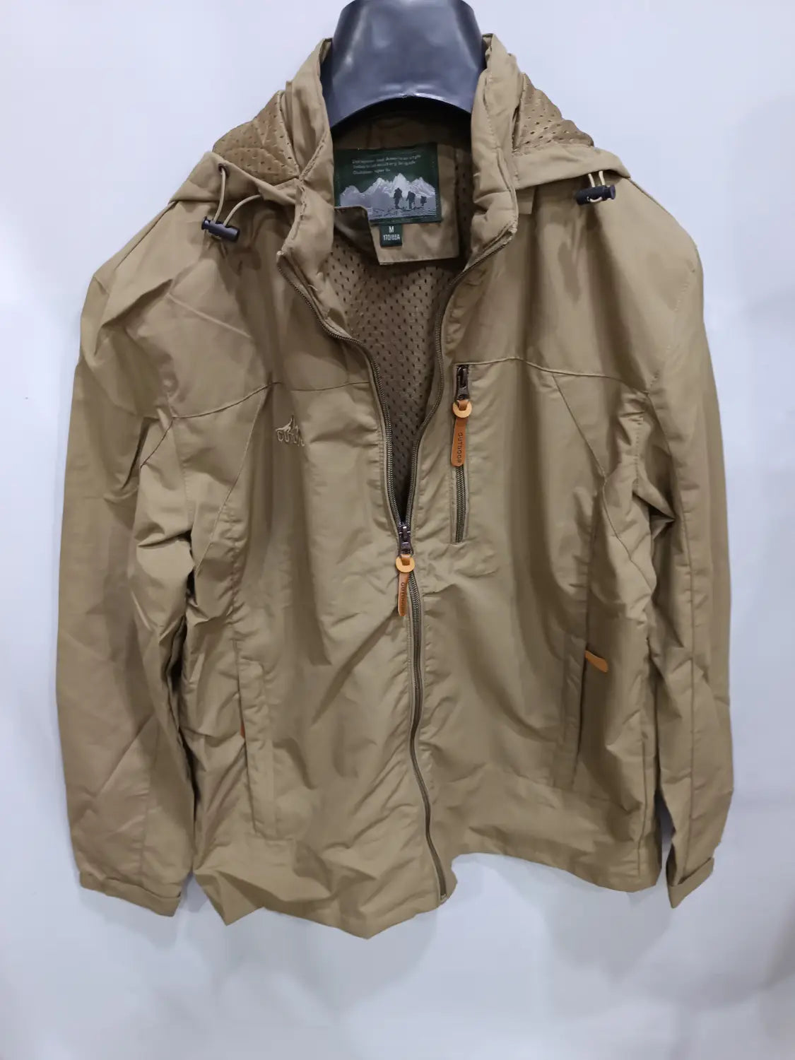 Chaqueta de senderismo con capucha impermeable color camel, con parte frontal con cremallera y bolsillos, con tiradores de cremallera de cuero color camel, diseñada para actividades al aire libre.