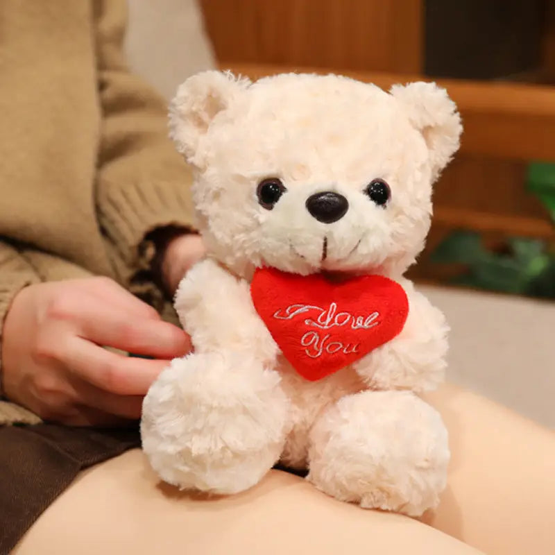 Ein weicher weißer Plüsch-Teddybär mit schwarzen Knopfaugen und einem angenähten Lächeln hält ein rotes Herz mit dem Text „I love you“; präsentiert als Teddybär-Plüschtier mit Rosen, Valentinsgeschenk, 16 bis 25 Zentimeter.