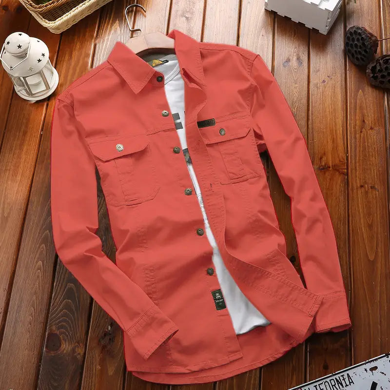 Chemise d’homme d’extérieur à manches longues unie de couleur corail, avec deux poches de poitrine et des boutons foncés, présentée sur un cintre en bois.
