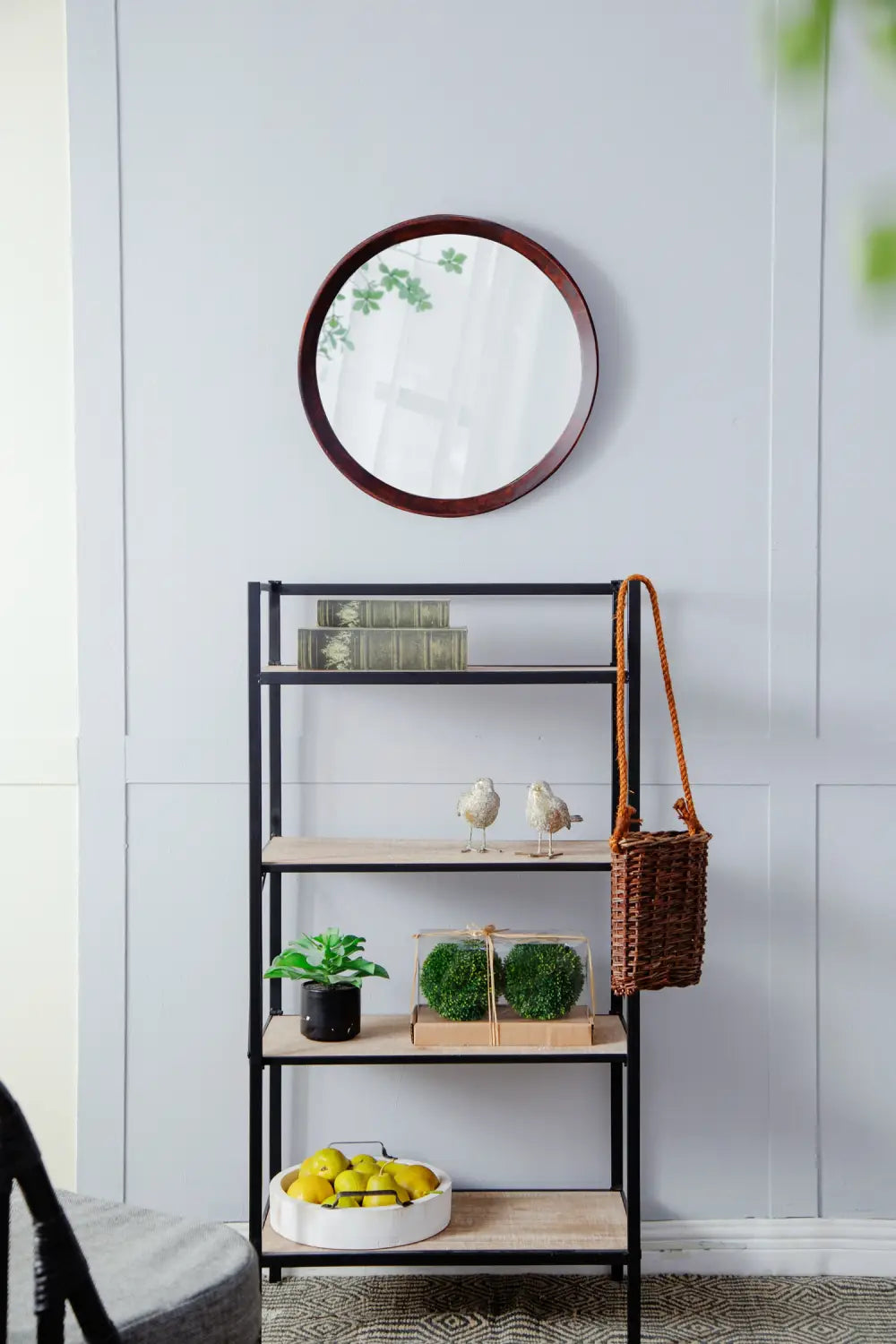 A mango wood round wall mirror with a 20 inch dark bronze frame hangs above a black metal and light wood shelf unit.
