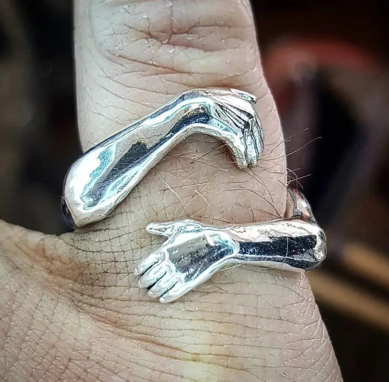 Adjustable silver ring featuring two intricately detailed hands clasping each other in a gentle embrace with a red gemstone.