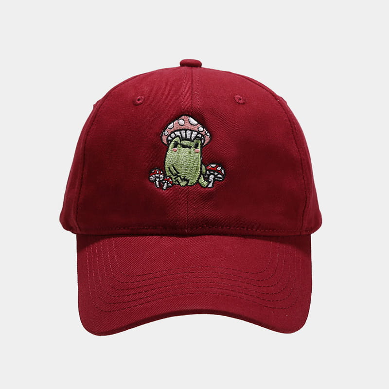 A maroon baseball cap featuring a charming embroidered design of a green frog sitting under a pink mushroom with white spots, surrounded by small red mushrooms in the frog mushroom embroidered baseball cap.